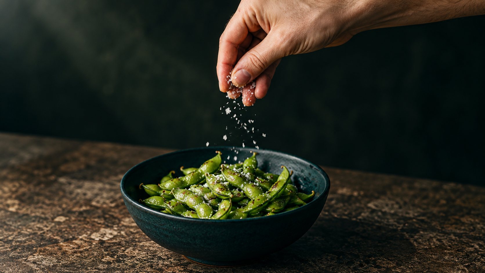 A hand sprinkling sea salt flakes over a bowl of grilled edamame